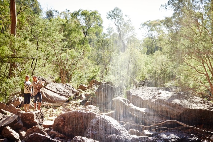 Two people standing on rocky terrain in a forest, with sunlight filtering through trees and a waterfall cascading in the background.