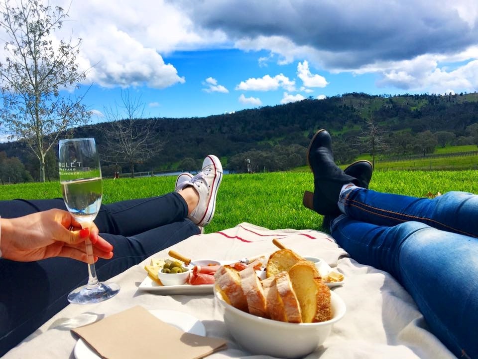 Two people lying on a blanket having a picnic with wine and food, set in a grassy area with rolling hills and a partly cloudy sky in the background.