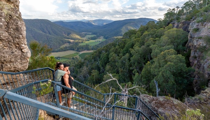 Two people standing on a metal staircase built along a rocky cliff, overlooking a lush green mountainous landscape with valleys under a partly cloudy sky.