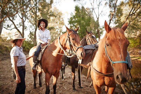 Girl sits on horse laughing in the Australian bush while lady watches on hand on horse